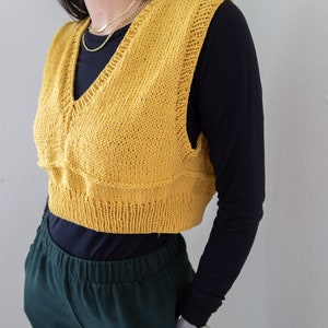 May include: Mustard yellow knitted vest with a V-neck. The cropped vest features a textured design on the front and a ribbed hem. The vest is worn over a black long-sleeved shirt and paired with green pants. A gold necklace is visible.