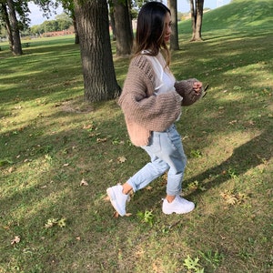 May include: A woman wearing a chunky knit, light brown cardigan over a white top and light blue jeans. She is wearing white trainers and walking on a grassy area with trees in the background.
