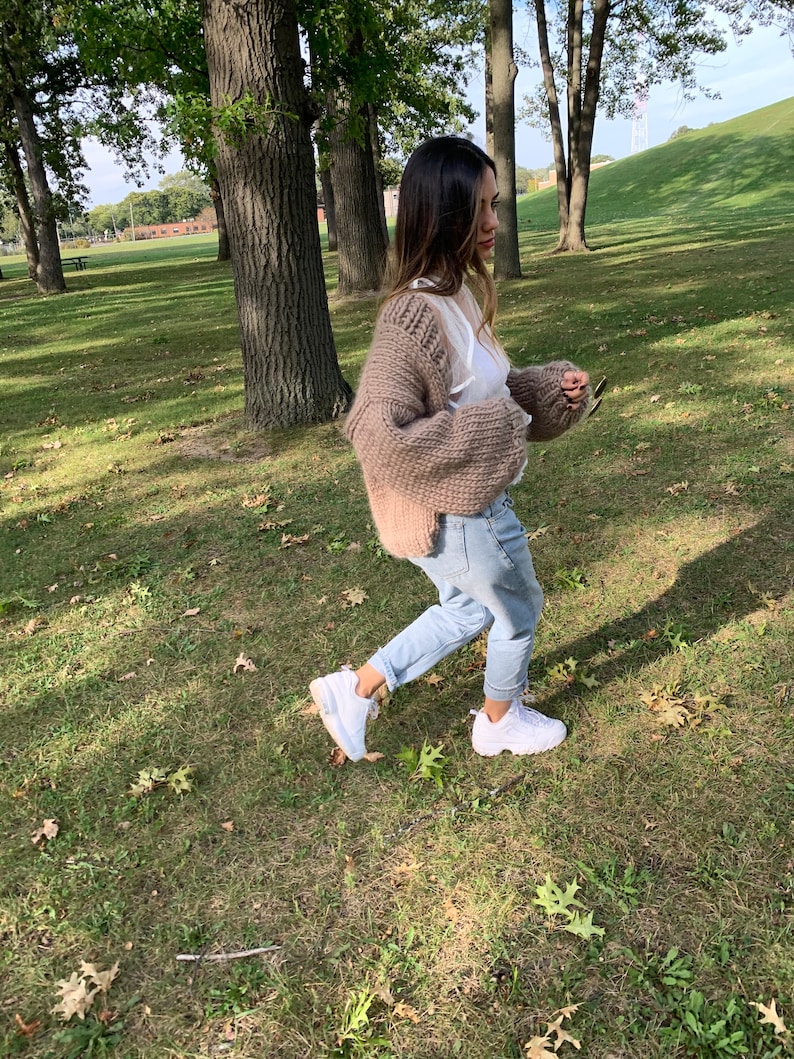 May include: A woman wearing a chunky knit, light brown cardigan over a white top and light blue jeans. She is wearing white trainers and walking on a grassy area with trees in the background.