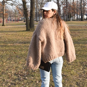 May include: A person wearing a beige, chunky knit jumper with long sleeves and light blue jeans. They are also wearing a white baseball cap and carrying a black purse. The background is a park with trees and grass.