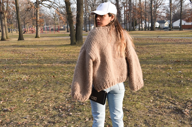 May include: A person wearing a beige, chunky knit jumper with long sleeves and light blue jeans. They are also wearing a white baseball cap and carrying a black purse. The background is a park with trees and grass.