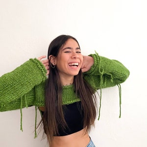 May include: A woman wearing a green knitted crop top with long sleeves and frayed edges. She is wearing blue jeans and has long brown hair.