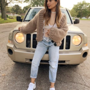 May include: A woman wearing a chunky knit, light brown cardigan with a white top, light blue jeans, and white trainers. The woman is standing in front of a gold-coloured car.
