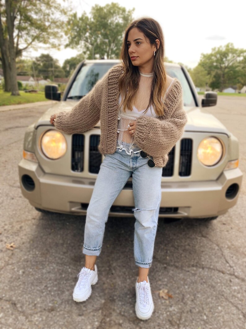 May include: A woman wearing a chunky knit, light brown cardigan with a white top, light blue jeans, and white trainers. The woman is standing in front of a gold-coloured car.