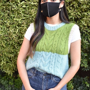 Op de afbeelding: Een zwart gezichtsmasker met een witte accentlijn wordt gedragen. Een groen en blauw kabelgebreid vest wordt over een wit t-shirt gedragen. De persoon draagt een blauwe spijkerbroek.
