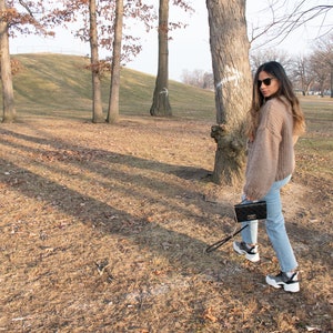 May include: A person wearing a tan knit sweater, light blue jeans, and black and white trainers, carrying a black clutch with a silver clasp. The person is walking in a park with trees and dry grass.