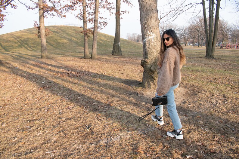May include: A person wearing a tan knit sweater, light blue jeans, and black and white trainers, carrying a black clutch with a silver clasp. The person is walking in a park with trees and dry grass.