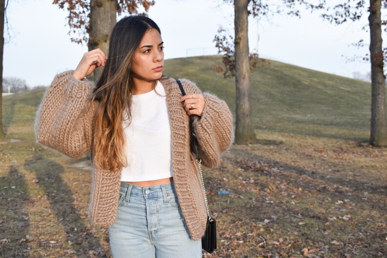 May include: A tan, open-front cardigan with oversized sleeves. The chunky knit sweater is paired with a white crop top and light wash jeans. A black purse is worn over the shoulder. The background includes trees and a grassy hill.