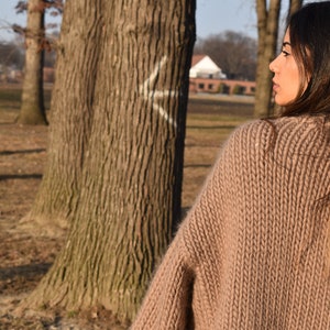 May include: A woman wearing a tan, chunky knit sweater outdoors. The sweater has a ribbed texture and a wide neckline. The background includes trees and a glimpse of a building.