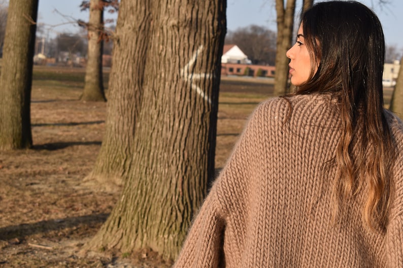 May include: A woman wearing a tan, chunky knit sweater outdoors. The sweater has a ribbed texture and a wide neckline. The background includes trees and a glimpse of a building.