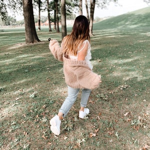 May include: A person wearing a light brown knit cardigan, a white top, light blue jeans, and white trainers, walking on a grassy area with trees in the background. The cardigan has long sleeves.