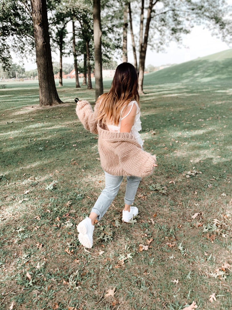 May include: A person wearing a light brown knit cardigan, a white top, light blue jeans, and white trainers, walking on a grassy area with trees in the background. The cardigan has long sleeves.