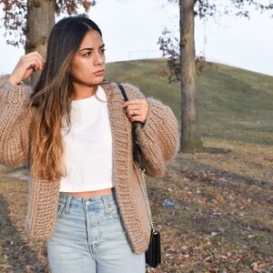 May include: A tan, open-front cardigan with oversized sleeves. The chunky knit sweater is paired with a white crop top and light wash jeans. A black purse is worn over the shoulder. The background includes trees and a grassy hill.