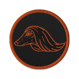 May include: Embroidered patch featuring a stylized dog head in profile. The design is rendered in black and outlined with orange thread, set against a black circular background. The patch has an orange border and is likely for apparel or accessories.