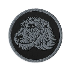Peut inclure: Écusson brodé représentant une tête de chien détaillée en nuances de gris. Le portrait du chien est centré sur un cercle noir, encadré d'une bordure grise. Le design présente des coutures complexes, créant un aspect texturé.