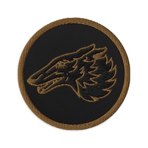 May include: Embroidered patch featuring a wolf's head profile in tan thread on a black circular background. The patch has a tan border. The wolf's head is detailed with intricate stitching, showcasing the animal's features.