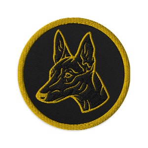 Könnte beinhalten: Gestickter Aufnäher mit einem schwarzen Hunde-Kopfprofil auf schwarzem, kreisförmigem Hintergrund. Die Konturen des Hundes sind gelb gestickt, und der Aufnäher hat einen gelben Rand. Das Design eignet sich für Kleidung oder Accessoires.