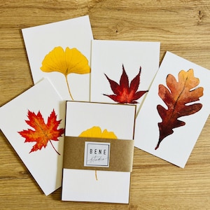 May include: A collection of watercolor greeting cards featuring autumn leaves in shades of yellow, red, and brown. The cards are arranged on a wooden surface, with a set of cards wrapped in a brown band. The band has the text "BENE studio".