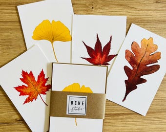 May include: A collection of watercolor greeting cards featuring autumn leaves in shades of yellow, red, and brown. The cards are arranged on a wooden surface, with a set of cards wrapped in a brown band. The band has the text "BENE studio".