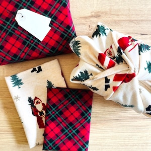May include: A collection of fabric items including a red and green plaid wrapped package with a blank white tag, a folded cream-colored fabric featuring Santa Claus and pine tree prints, and a red and green plaid fabric square.