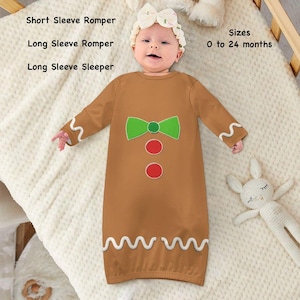 Op de afbeelding: Een bruine baby slaapzak met lange mouwen met een peperkoekmannetje ontwerp, met een groene strik en rode knopen. De slaapzak heeft witte zigzag trim en is verkrijgbaar in maten 0 tot 24 maanden. De tekst toont "Short Sleeve Romper", "Long Sleeve Romper", en "Long Sleeve Sleeper".