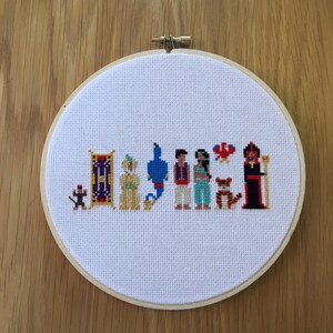 May include: Cross-stitch embroidery hoop with a scene from the Disney movie Aladdin. The characters include Aladdin, Jasmine, Genie, Abu, Jafar, and Rajah. The characters are all in their iconic outfits and poses.