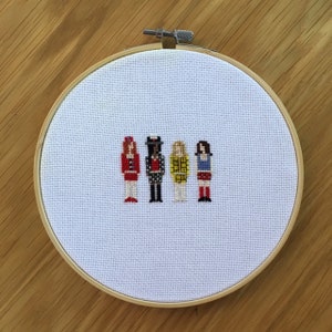 May include: Cross-stitch embroidery hoop with a white fabric background featuring five pixelated figures in a row. The figures are dressed in red, black and white, yellow, and blue.