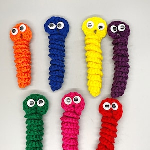 May include: Seven handmade crochet worms in various colors: orange, blue, yellow, purple, green, pink, and red. Each worm has googly eyes and a textured, segmented body. The worms are arranged on a light gray surface.