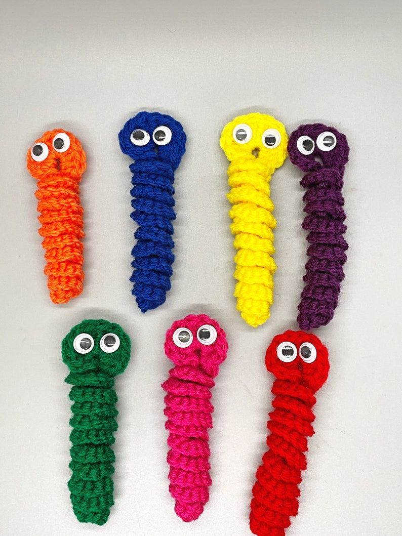 May include: Seven handmade crochet worms in various colors: orange, blue, yellow, purple, green, pink, and red. Each worm has googly eyes and a textured, segmented body. The worms are arranged on a light gray surface.