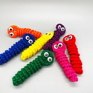 May include: A collection of colorful, crocheted worm toys. Each worm is a different vibrant color, including orange, blue, yellow, purple, pink, red, and green. They have googly eyes attached, giving them a playful appearance. The worms are arranged on a plain white background.
