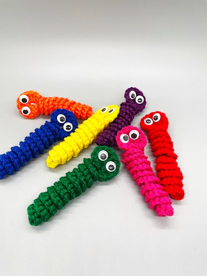 May include: A collection of colorful, crocheted worm toys. Each worm is a different vibrant color, including orange, blue, yellow, purple, pink, red, and green. They have googly eyes attached, giving them a playful appearance. The worms are arranged on a plain white background.