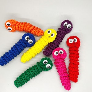 May include: A collection of handmade crochet worms in various bright colors, including orange, blue, yellow, green, purple, pink, and red. Each worm has googly eyes attached, giving them a playful appearance. The worms are arranged on a light gray surface.