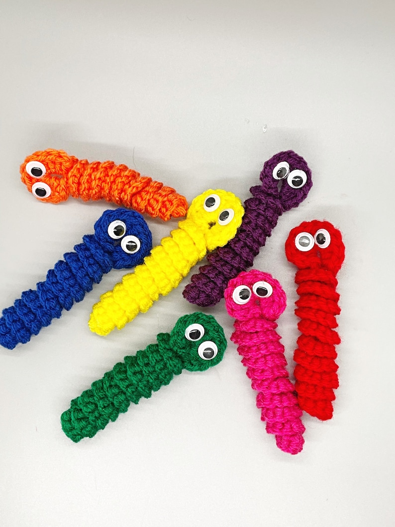 May include: A collection of handmade crochet worms in various bright colors, including orange, blue, yellow, green, purple, pink, and red. Each worm has googly eyes attached, giving them a playful appearance. The worms are arranged on a light gray surface.