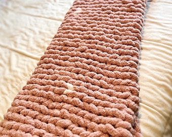 May include: A soft, pink knitted throw blanket with a chunky knit texture. The blanket is folded and lying on a white bed.