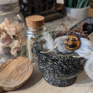 May include: A grey stone mortar and pestle with a small, handmade scarecrow figure inside. The scarecrow has a brown burlap hat, a white fuzzy face, and a painted orange pumpkin face with a smiling expression.