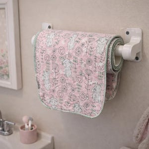 May include: A pink and white patterned paper towel roll with a bunny and floral design, mounted on a white holder. The roll is displayed in a bathroom setting, with a framed bunny picture and a plush bunny toy.