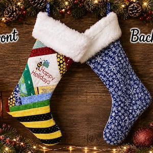 May include: A Christmas stocking with two sides shown. The front features a patchwork design of yellow, green, and blue fabrics with a bee graphic and the text Happy Holidays. The back is blue with a white snowflake pattern. Both have white cuffs.