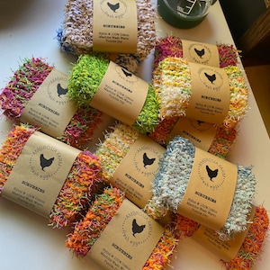 May include: Nine colourful crocheted scrubbies with a brown label that reads "Ambrosia Farm Fresh Eggs Scrubbie Nylon & 100% Cotton Machine Wash Warm Tumble Dry"
