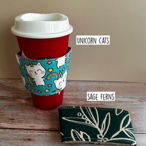 May include: A red reusable cup with a white lid and a teal fabric sleeve featuring a pattern of white unicorn cats, rainbows, and stars. Next to it lies a folded dark green fabric sleeve with a white fern leaf pattern labelled SAGE FERNS.