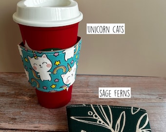 May include: A red reusable cup with a white lid and a teal fabric sleeve featuring a pattern of white unicorn cats, rainbows, and stars. Next to it lies a folded dark green fabric sleeve with a white fern leaf pattern labeled SAGE FERNS.