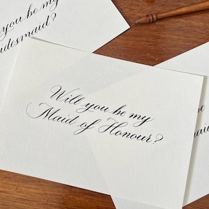 May include: Ivory cards with black calligraphy. The cards feature the phrases "Will you be my Maid of Honour?" and "Will you be my bridesmaid?" A wooden pen rests on the wooden surface.