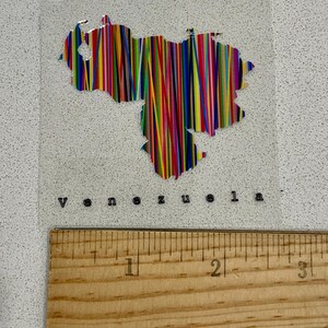 May include: A clear sticker featuring the outline of Venezuela filled with vertical stripes in various colors. The word "venezuela" is printed below the map. A wooden ruler is at the bottom.