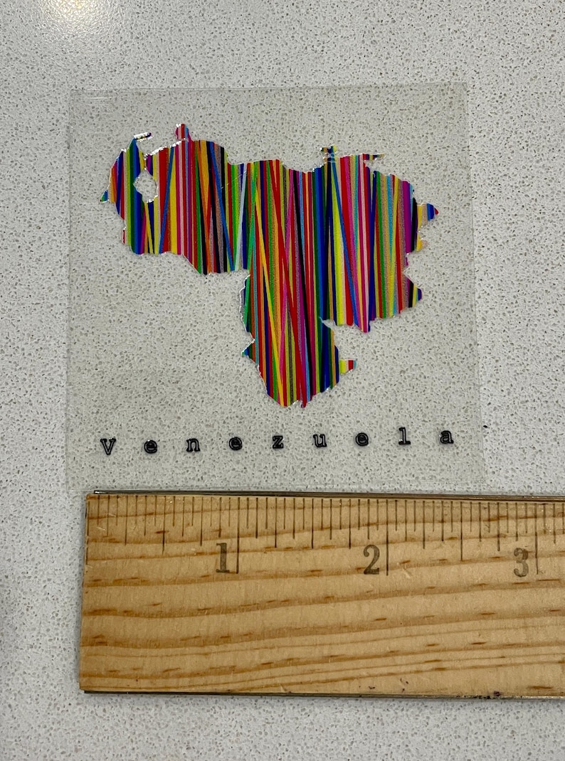 May include: A clear sticker featuring the outline of Venezuela filled with vertical stripes in various colors. The word "venezuela" is printed below the map. A wooden ruler is at the bottom.