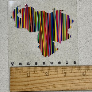 May include: A clear, rectangular sticker featuring the outline of Venezuela filled with vertical stripes in various colors. The word "Venezuela" is printed below the image. A wooden ruler is placed below the sticker.