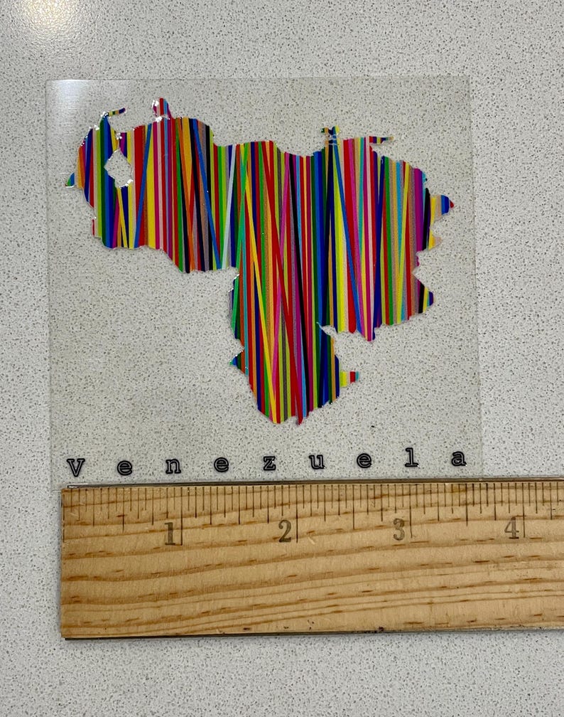 May include: A clear, rectangular sticker featuring the outline of Venezuela filled with vertical stripes in various colors. The word "Venezuela" is printed below the image. A wooden ruler is placed below the sticker.