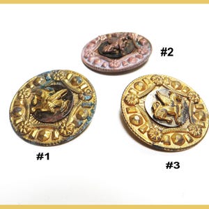 May include: Three antique buttons with detailed designs. Button #1 is oval, gold-toned with a central emblem and floral accents. Button #2 is oval, with a pinkish-brown hue and a central figure. Button #3 is round, gold-toned with a winged creature.