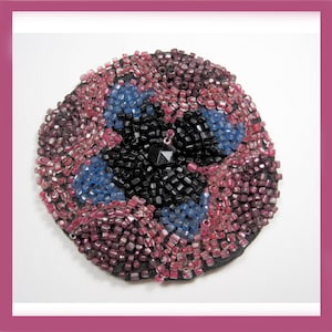 May include: Round beaded brooch with a floral design. The brooch features a black center, blue petals, and pink and purple beads around the edges. A small, black, faceted gem is in the center.