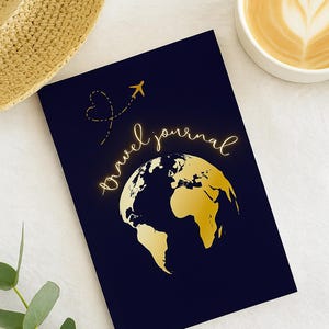 May include: A navy blue travel journal featuring a gold world map and the words "travel journal" in gold script. A gold airplane graphic and heart shape are above the text. A cup of coffee and a straw hat are in the background.