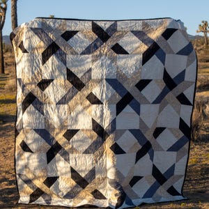 May include: A geometric quilt with a black, beige, and blue pattern. The quilt is displayed outdoors, with a black border and a design featuring intersecting shapes. The background shows a desert landscape with Joshua trees.