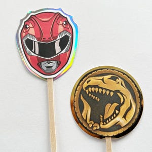 Puede incluir: Dos adornos para cupcakes con tem&aacute;tica de Power Rangers. Uno presenta un casco de Ranger rojo con un contorno plateado. El otro es una moneda de oro con un dise&ntilde;o de dinosaurio negro. Ambos est&aacute;n unidos a palitos de madera.